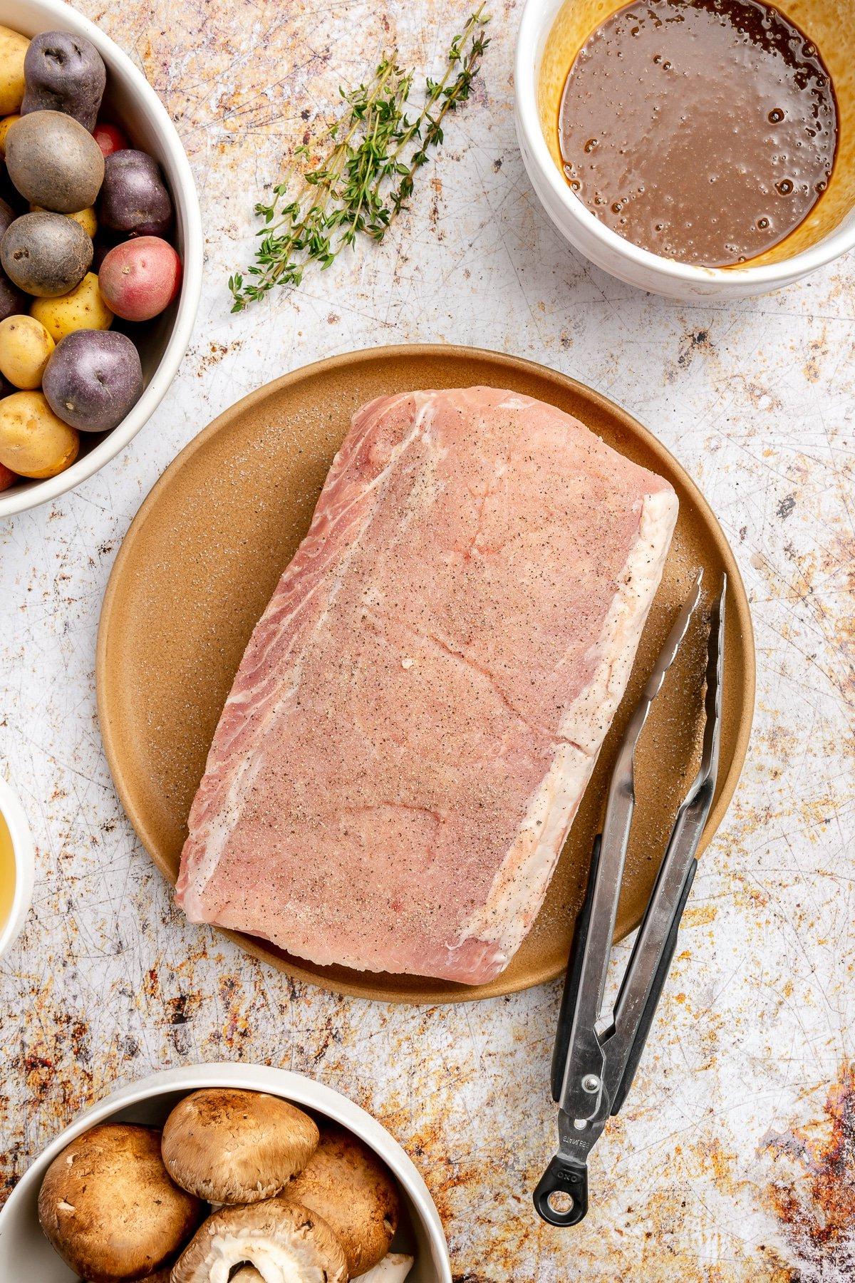 Die Gewürzmischung wurde auf die Oberfläche eines Schweinefilets gerieben, das auf einem braunen Teller liegt. Eine schwarze Zange liegt daneben.