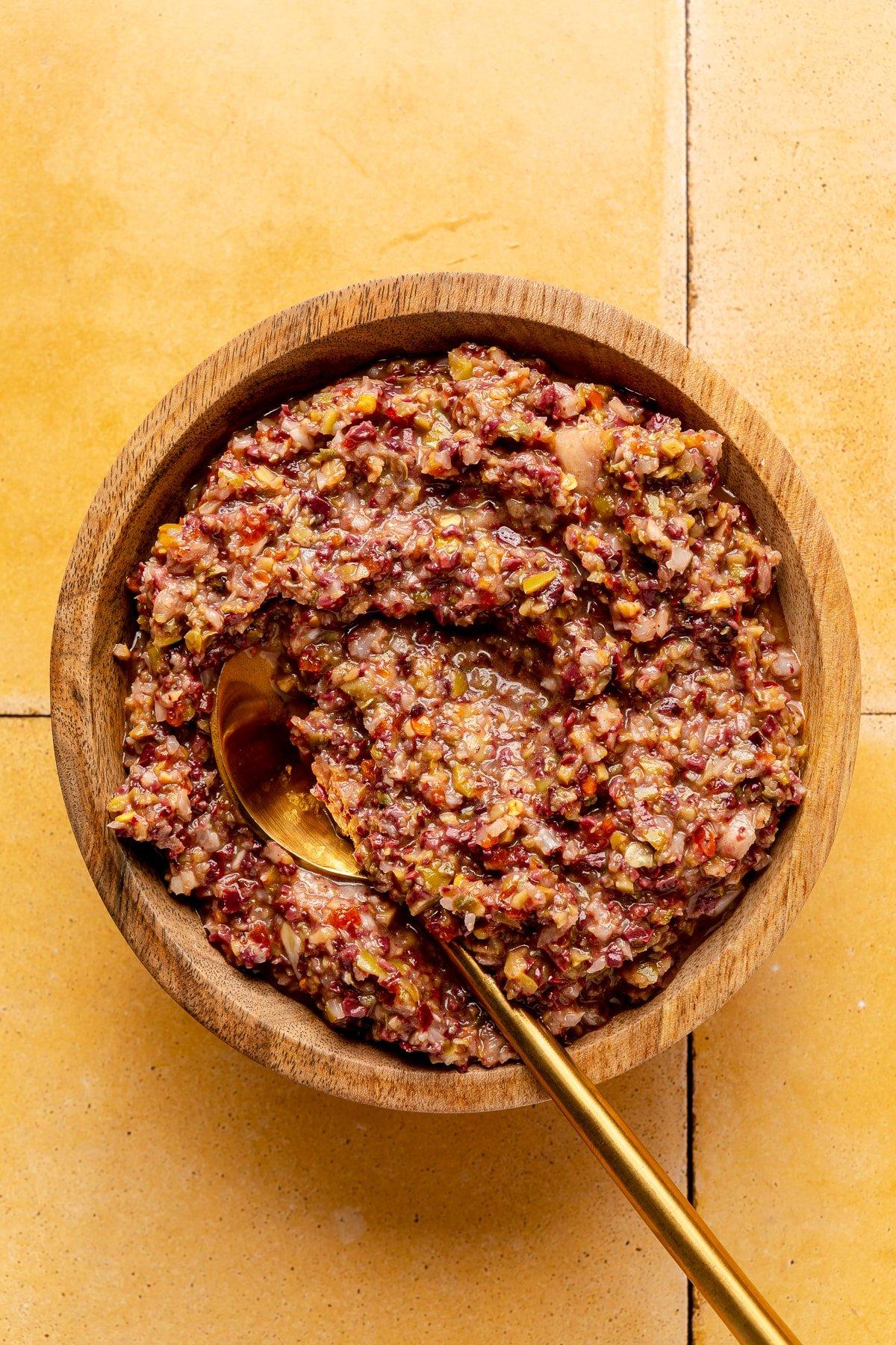 Oliven-Tapenade in einer hölzernen Servierschale. Ein goldfarbener Löffel liegt daneben.