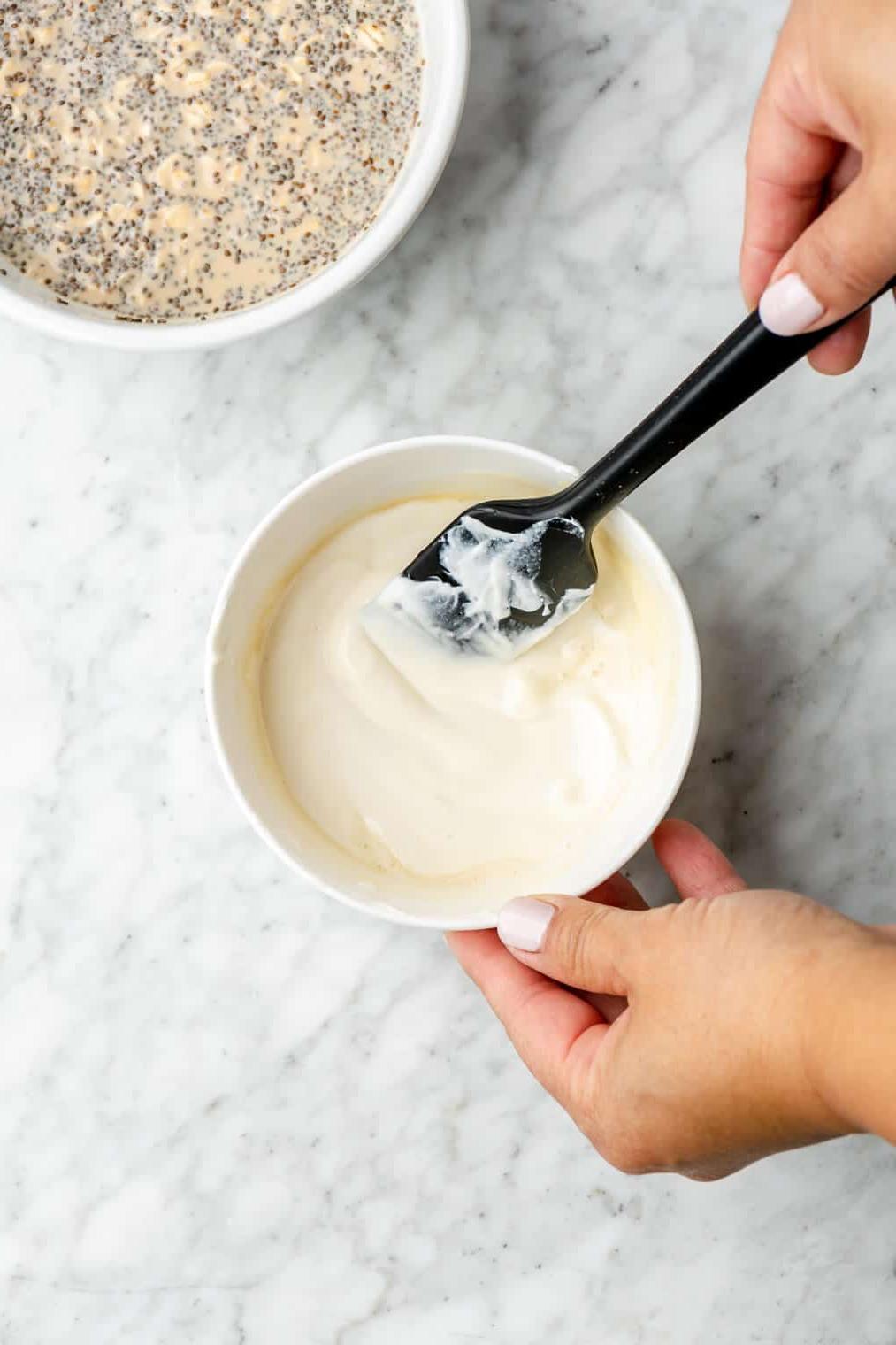 Hand rührt mit einem schwarzen Spatel Joghurt und Ahornsirup in einer kleinen weißen Schüssel zusammen.