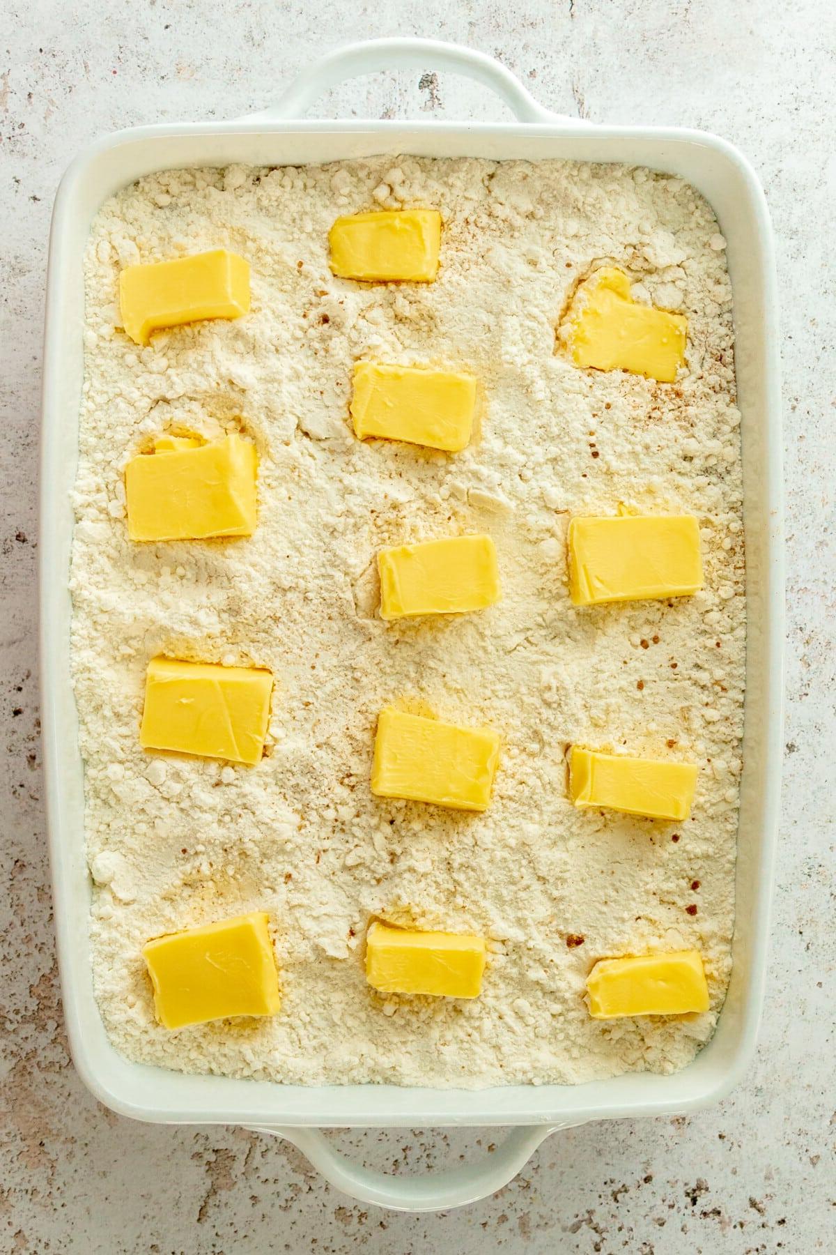 Butterscheiben liegen auf einer Kürbis-Dump-Cake-Mischung in einer weißen, rechteckigen Auflaufform auf hellgrauem Untergrund.