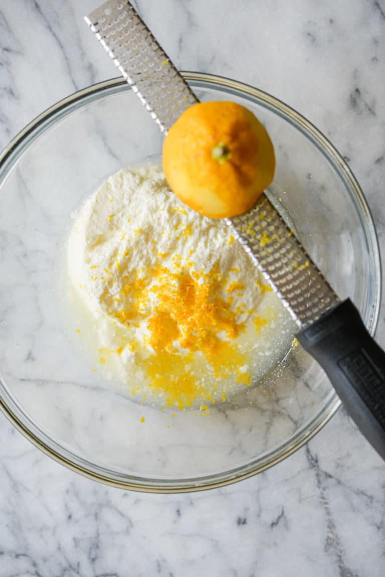 Joghurt, Zitronensaft und Zitronenschale in einer durchsichtigen Schüssel mit einem Zestenreißer und einer halbierten Zitrone auf dem Schüsselrand