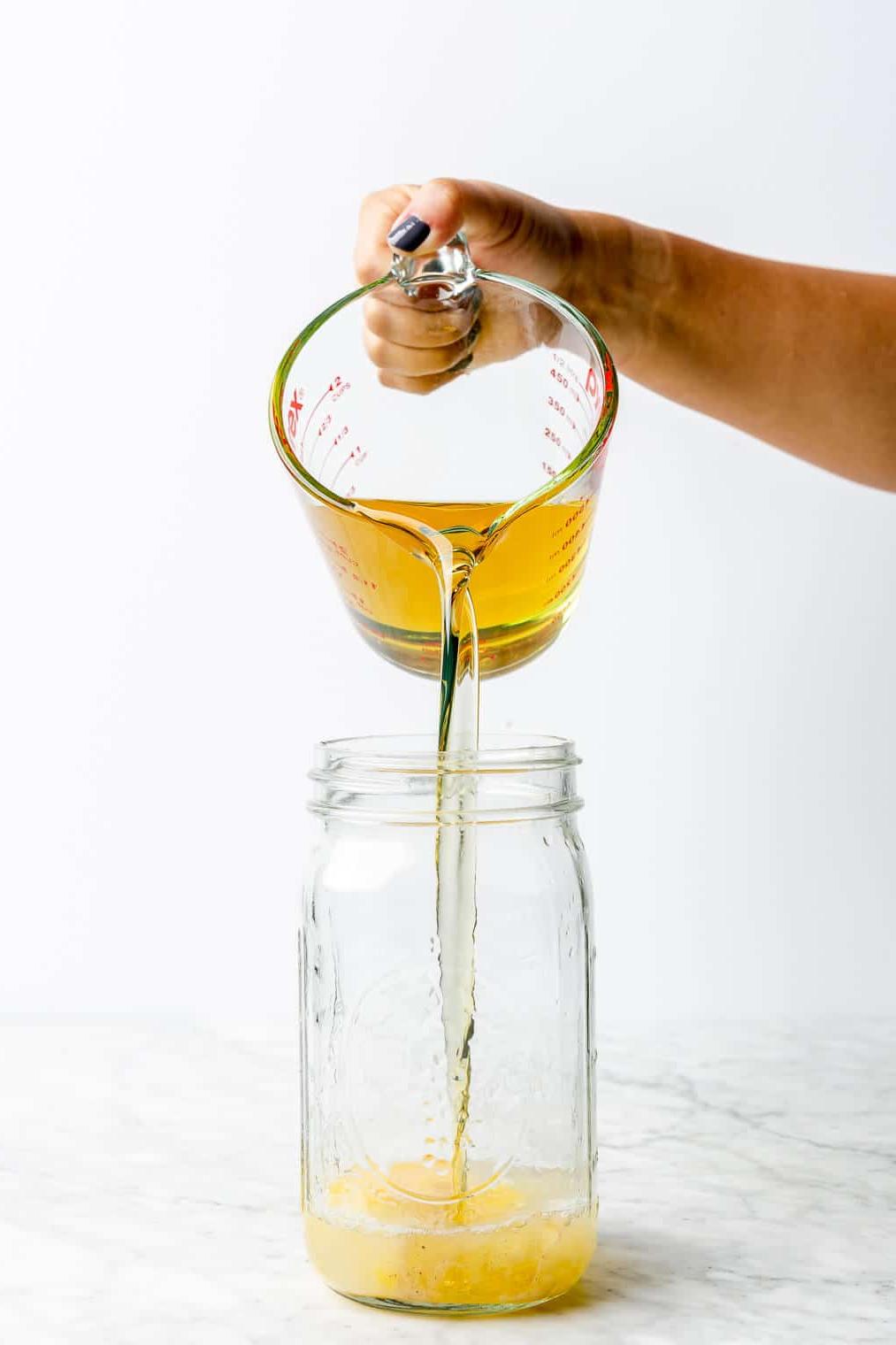 Hand, die Öl in ein großes Einmachglas mit aufgeschlagenem Ei und Zitronensaft auf einer grau-weißen Marmoroberfläche gießt.