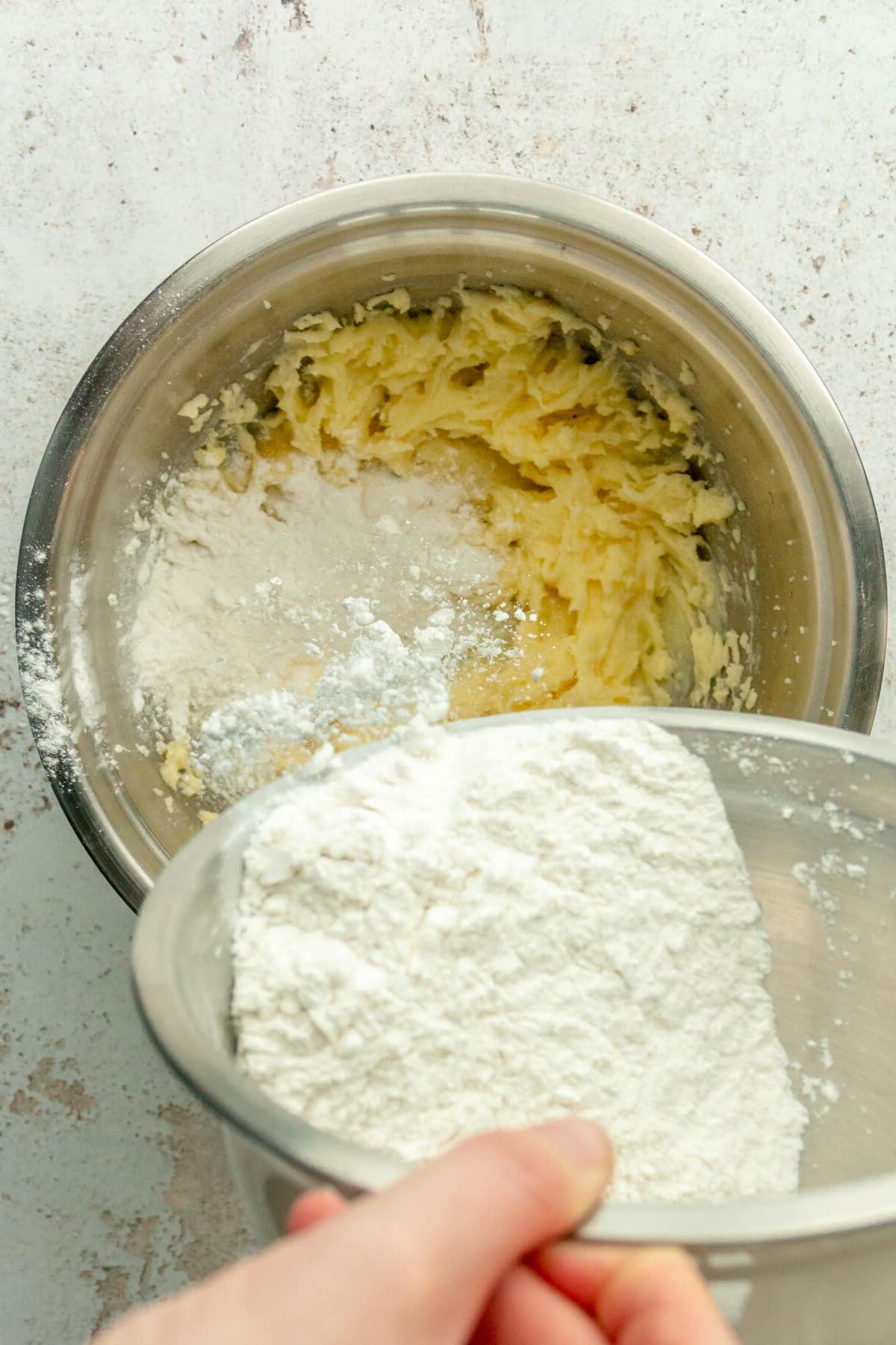 Eine Mischung aus Mehl und Triebmitteln wird in eine geschlagene Butter-Zucker-Mischung in einer Edelstahlschüssel auf hellem grauem Untergrund gegeben.