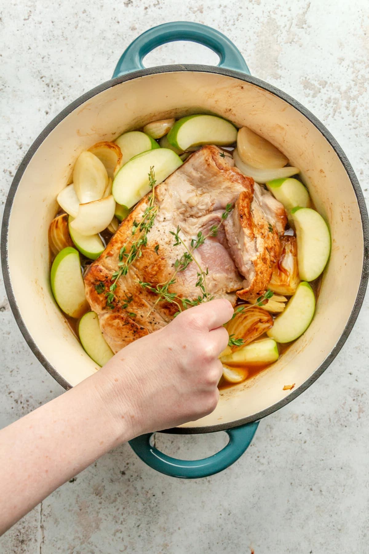 Frischer Thymian wird auf Schweinefilet gelegt, umgeben von geviertelten Äpfeln und Zwiebeln in einem Bräter auf einer hellgrauen Oberfläche.