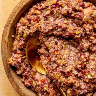 Oliven-Tapenade in einer hölzernen Servierschale. Ein goldfarbener Löffel liegt daneben.