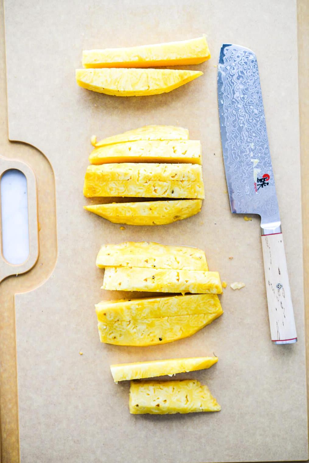 Ein großes Schneidebrett mit Streifen frisch geschnittener Ananas liegt neben einem großen Messer.