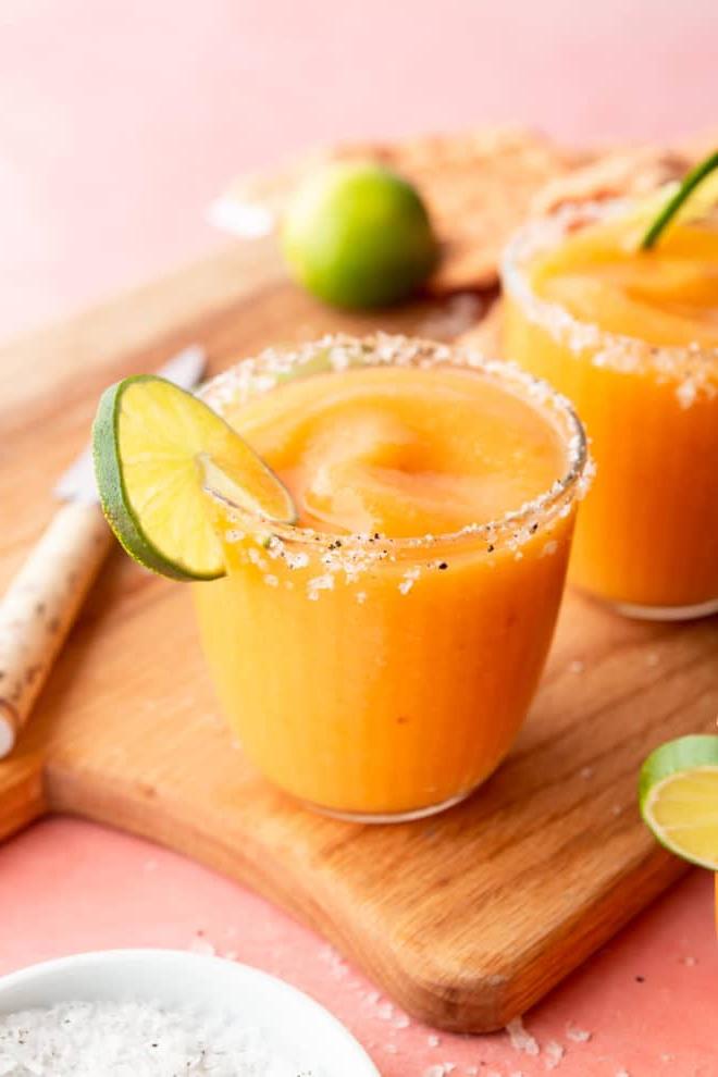 Seitenansicht von gefrorenen Pfirsich Margaritas in Gläsern, garniert mit Meersalzflocken und einer Limettenscheibe, auf einer pinkfarbenen Oberfläche mit Schneidebrett, Schälmesser, einer Prise Meersalzflocken, Limettenscheiben und orangefarbenem Leinentuch.