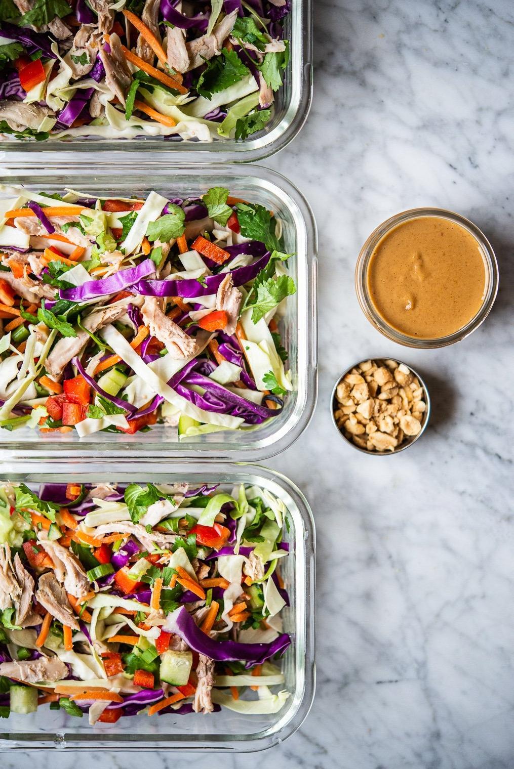 Ein farbenfroher Thai-Hähnchensalat in 3 einzelnen Glas-Meal-Prep-Behältern neben Erdnussdressing und zerstoßenen Erdnüssen