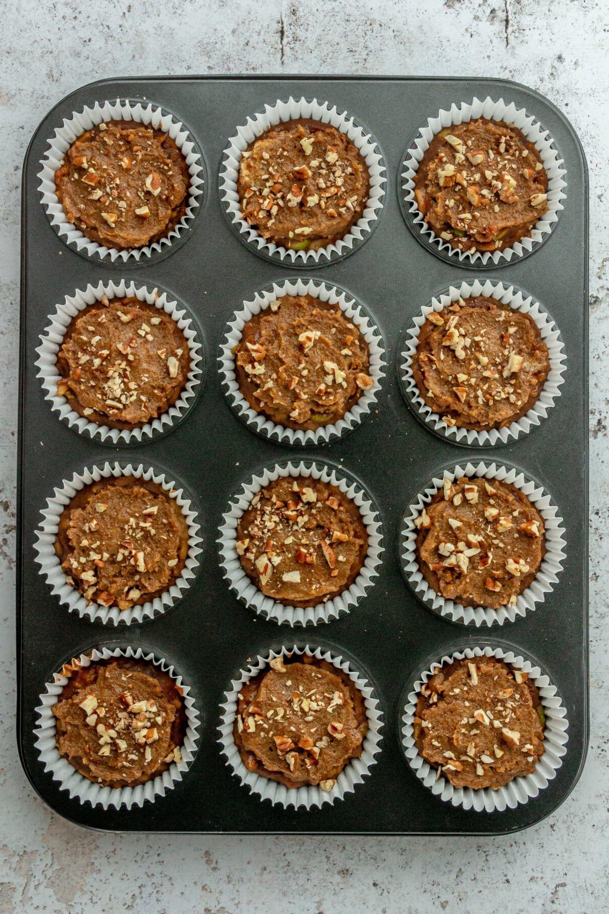 Paleo apple cinnamon muffin batter topped with a crumble mixture and pecans batter sits in white liners in a 12 muffin tin on a light grey surface.