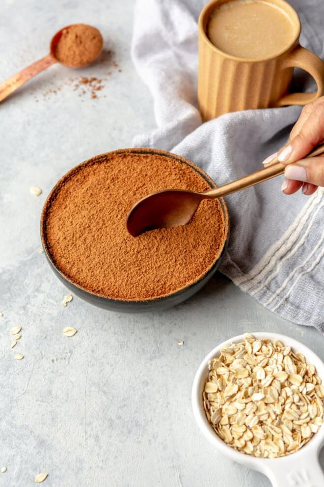 Hand hält einen Löffel und bereitet sich darauf vor, eine Portion Overnight Oats aus einer Schale zu nehmen. Auf dem Tisch stehen außerdem eine Tasse Kaffee, ein Messlöffel mit Kakaopulver und ein Messbecher mit trockenen Haferflocken.