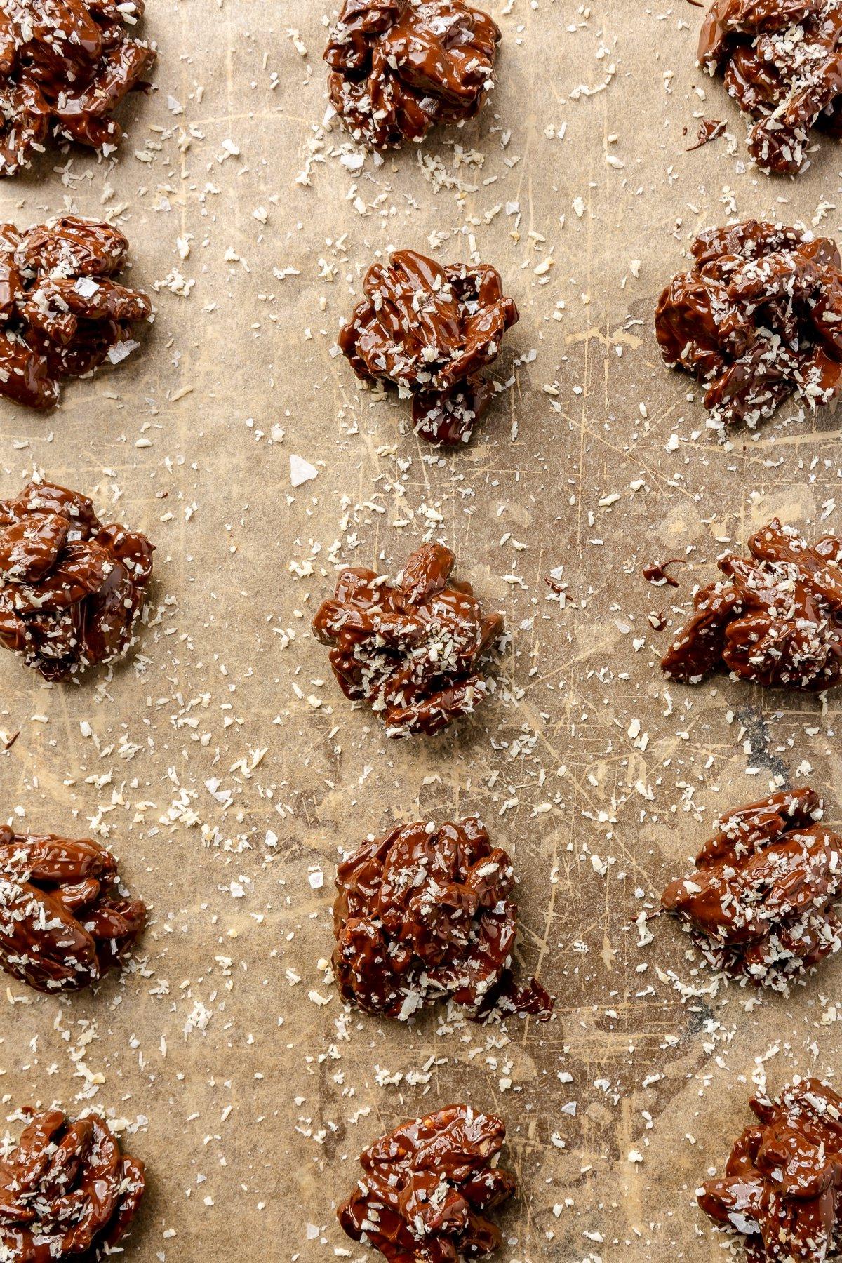 Dunkle Schokolade-Chili-Pekannuss-Brocken 8 Frisch geformte Schoko-Chili-Pekannuss-Häufchen auf Backpapier, bereit zum Bestreuen mit Kokosraspeln und Meersalz.