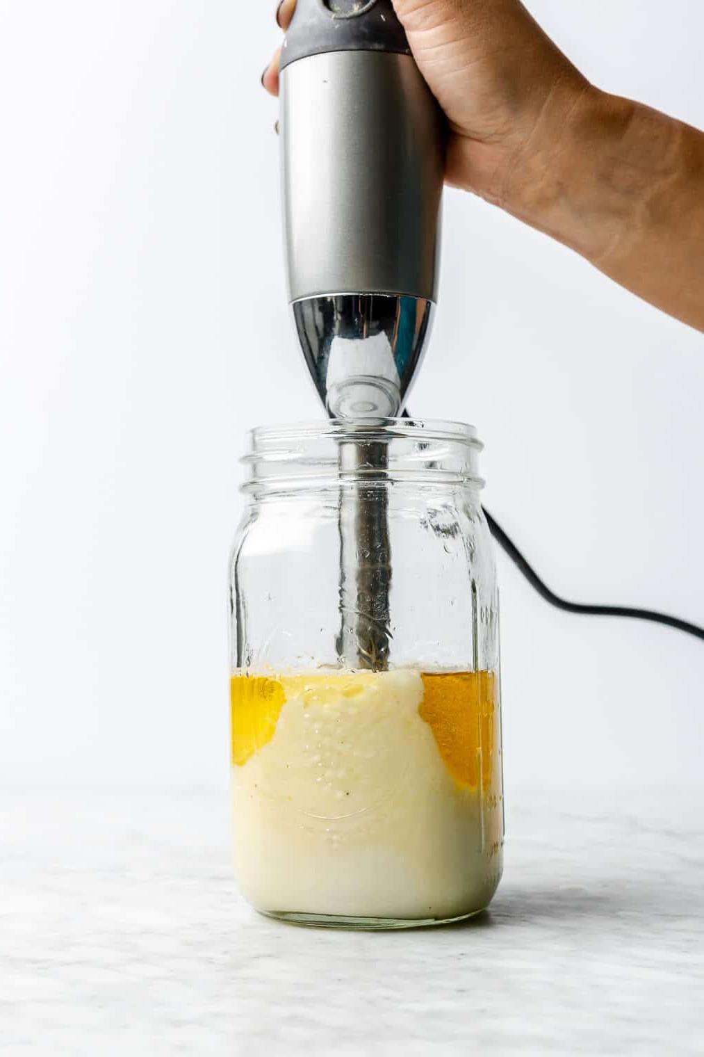 Hand, die einen Stabmixer in einem großen Einmachglas hält und die hausgemachte Mayonnaise püriert.