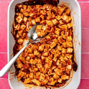 Gebackene BBQ-Hähnchenbrüste gewürfelt in einer großen weißen Auflaufform auf einem pinkfarbenen Fliesenuntergrund.
