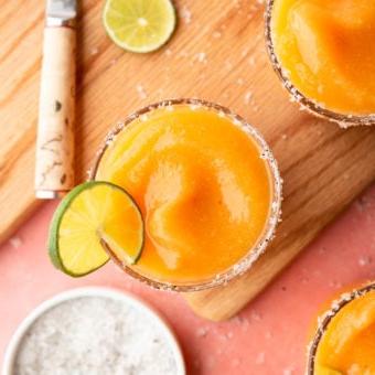 Aufnahme von oben: Gefrorene Pfirsich Margaritas in Gläsern, garniert mit Meersalzflocken und einer Limettenscheibe, auf einer pinkfarbenen Oberfläche mit Schneidebrett, Schälmesser, einer Prise Meersalzflocken, Limettenscheiben und orangefarbenem Leinentuch.