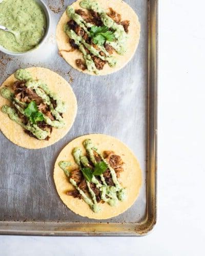 210414_Carnitas-35 3 offene Carnitas Tacos, belegt mit cremiger Avocado-Verde-Sauce und Koriander, auf einem Backblech neben einer Schüssel cremiger Avocado-Verde-Sauce