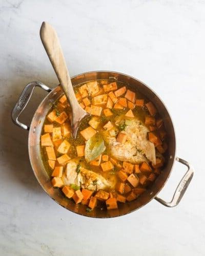 Ein Topf mit Brühe, gewürfelten Süßkartoffeln und Hähnchenbrust mit einem Holzlöffel darin