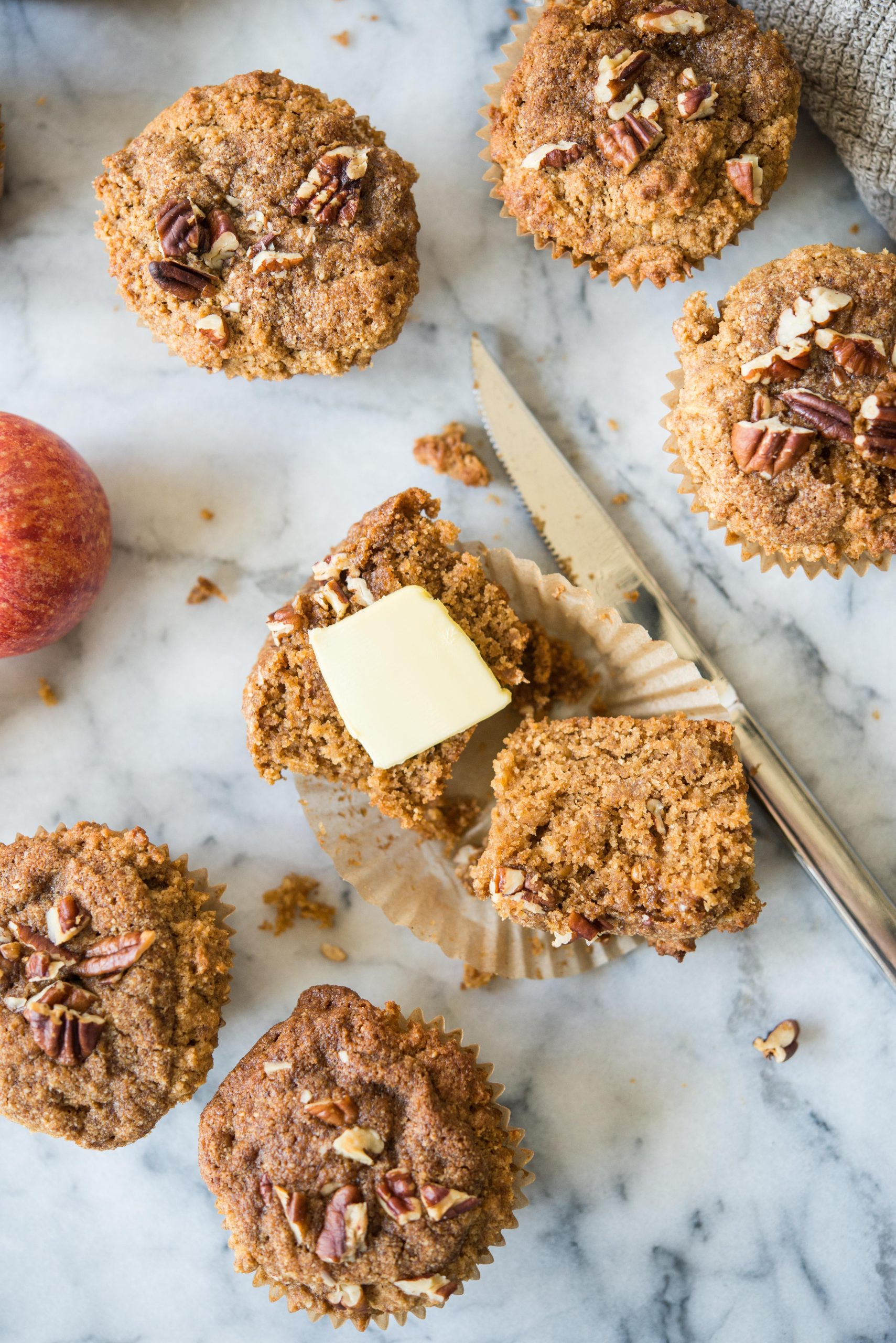 an apple cinnamon muffin cut in half with butter on top surrounded by more muffins all on a marble countertop