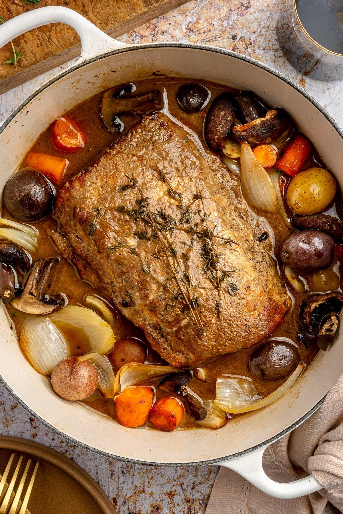 Ein fertig gegarter Balsamico-Schweinebraten liegt in einem weißen, emaillierten Topf. Zwei Weingläser, ein brauner Teller mit goldenem Besteck und ein Schneidebrett stehen daneben.