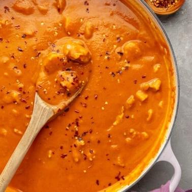 Einfaches Kürbiscurry-Rezept 9 Ein weißer Topf mit Kürbis-Curry, das mit roten Pfefferflocken bestreut ist. Ein Holzlöffel ist zu sehen, wie er einen Teil des Currys schöpft.