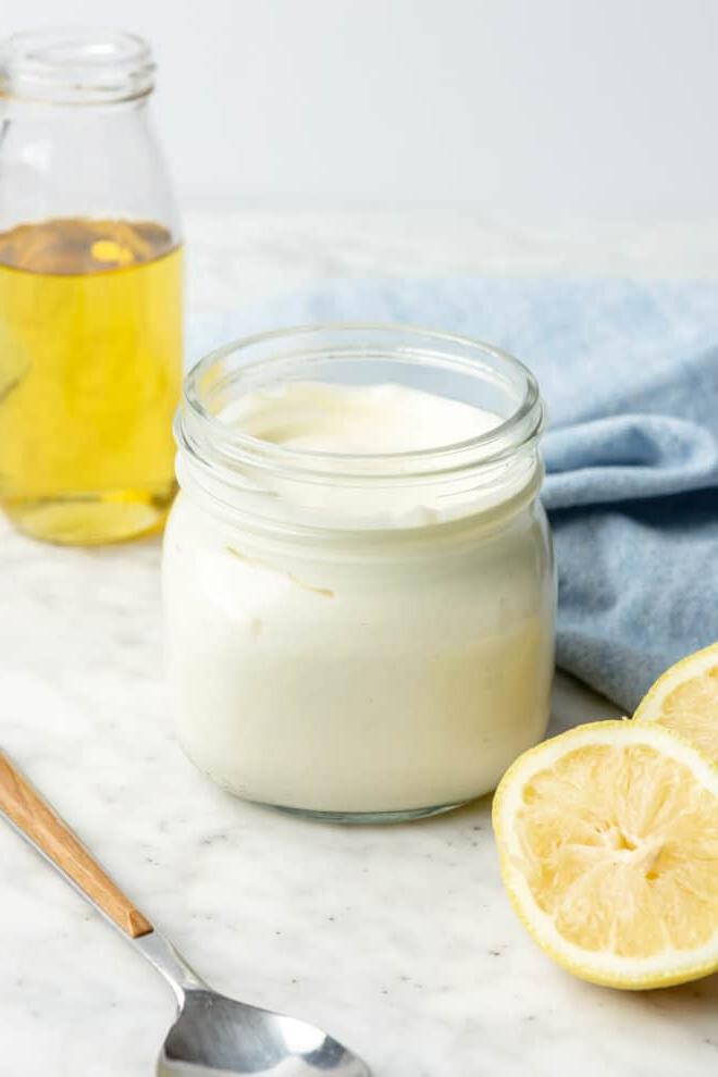 Glas mit hausgemachter Mayonnaise auf einer grau-weißen Theke mit Zitronenhälften, Avocadoöl im Hintergrund, einem Servierlöffel und einem hellblauen Leinentuch.