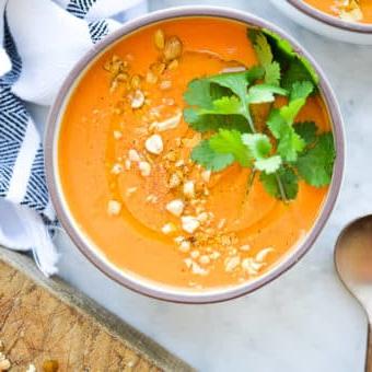 Eine Schüssel mit leuchtend oranger Thai-Karottensuppe, garniert mit frischem Koriander und zerdrückten Erdnüssen auf einer Marmoroberfläche.