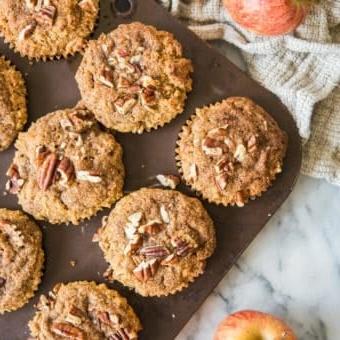 paleo apple cinnamon muffins in a muffin tin on a marble board with apples and a grey cloth surrounding them